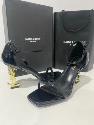 Svarta Saint Laurent pumps med YSL-klack - Snygga svarta pumps från Saint Laurent i lackat läder med öppen tå och smal rem över foten. Klacken är i guld med ikoniska YSL-loggan som design. Fyrkantig tå och justerbar rem runt ankeln ger en modern och lyxig look.