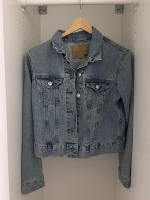 Klassisk ljusblå jeansjacka - Snygg ljusblå jeansjacka med raka linjer och silverfärgade knappar. Jackan har två bröstfickor med lock och knapp samt klassisk krage. Perfekt att slänga över en hoodie eller t-shirt för en avslappnad look.