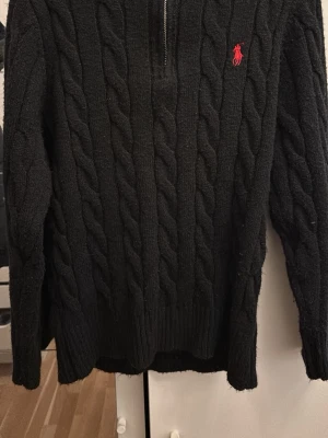 Svart kabelstickad tröja Polo Ralph Lauren - Svart kabelstickad tröja från Polo Ralph Lauren med halv dragkedja och röd broderad logga på bröstet. Tröjan har ribbstickade muddar och klassisk passform. Perfekt för en stilren och avslappnad look.
