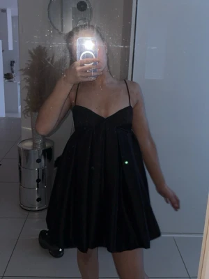 Svart kort klänning med tunna axelband - En svart, kort klänning med tunna axelband och v-ringad byst. Klänningen har en lätt utsvängd passform och är tillverkad i ett glansigt tyg som ger en elegant look. Perfekt för dig som gillar stilrena och trendiga plagg.