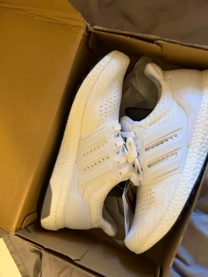 Vita Adidas Ultraboost  - Fräscha vita Adidas Ultraboost sneakers med stickad ovandel och klassiska tre ränder på sidan. Skorna har en tjock, responsiv Boost-sula och svart yttersula för extra grepp. Perfekta för dig som gillar stilrena och bekväma sneakers med modern design.