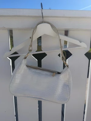 Vit axelväska i croco-mönster - Snygg vit handväska med croco-mönstrad yta och guldfärgade detaljer. Väskan har en kort axelrem och stängs med dragkedja. Perfekt för dig som gillar trendiga accessoarer med stilren look. Väskan är något smutsig men är annars i fint skick