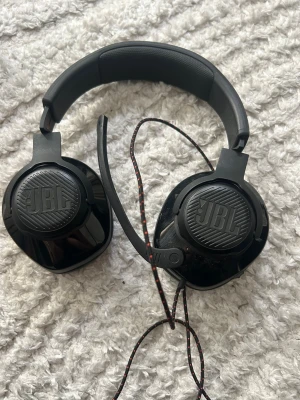 Svarta JBL over-ear hörlurar med sladd - Säljer ett par svarta over-ear hörlurar från JBL med kabel och mikrofon. Hörlurarna har en robust design med vadderad bygel och rundade kåpor. Sladden är svart med röda detaljer. Perfekt för gaming eller musik.