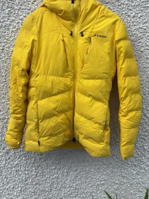 Gul dunjacka från Everest med huva - Snygg gul dunjacka från Everest med huva och dragkedja framtill. Jackan har flera praktiska fickor med dragkedja och en sportig look. Perfekt för kalla vinterdagar när du vill sticka ut i mängden.