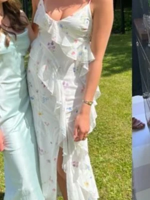 Vit långklänning med volanger och blommor - Superfin vit långklänning med tunna axelband och volangdetaljer. Klänningen har ett blommigt mönster i pastellfärger och en lätt, flowy känsla. Perfekt för sommaren!