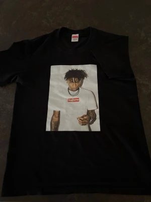 Supreme svart t-shirt med tryck - Svart t-shirt från Supreme med stort tryck på framsidan av en person i vit Supreme t-shirt. Klassisk rund hals och korta ärmar. Perfekt för dig som gillar streetwear och vill sticka ut med en ikonisk design.