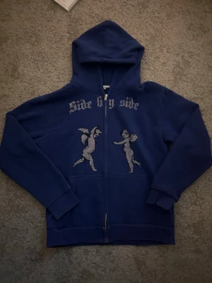 Blå hoodie med strasstenar och änglar - Mörkblå hoodie med dragkedja och huva. På bröstet står det 'side by side' i strasstenar och under finns två änglar i samma stil. Hoodien har ribbade muddar och är i mjukt material, perfekt för en chill och trendig look. Storlek XS men är stor i storlek så den kan passa om man har storlek S