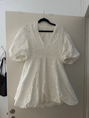 Vit kortklänning med puffärmar (studentklänning) - Supersöt vit kortklänning med stora puffärmar och v-ringning. Klänningen har ett blommigt broderat mönster och ballongformad kjol. Perfekt för studenten, säljer pågrund av att den var lite för liten. Står storlek L men skulle mer säga M 