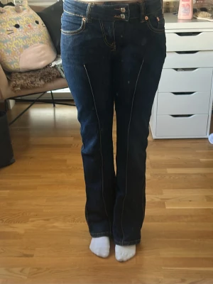 Mörkblå bootcut jeans med kontrastsömmar - Säljer ett par mörkblå bootcut jeans med snygga kontrastsömmar längs benen och klassiska fem fickor. Jeansen har hög midja och stängs med knapp och dragkedja. Perfekta för dig som gillar en retroinspirerad look.