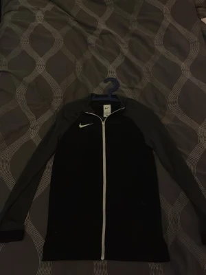 Svart träningsjacka från Nike - Svart träningsjacka från Nike med grå ärmar och hel dragkedja framtill. Jackan har lång ärm, vit Nike-logga på bröstet och är tillverkad i ett lätt och smidigt material som passar perfekt till träning.