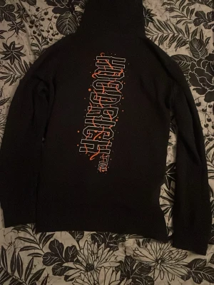 Svart hoodie från Hoodrich - Säljer en svart hoodie från Hoodrich med stor logga i vitt och orange på bröstet och en vertikal grafisk text på ryggen. Hoodien har huva och en mindre tryckt detalj på magen. Perfekt för dig som gillar streetwear och vill sticka ut.