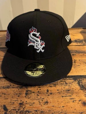 Svart New Era White Sox keps - Säljer en svart New Era 59FIFTY keps med White Sox-logga framtill i vitt med röda och blåa flammor. Kepsen har broderade detaljer på sidorna och baksidan, samt en rak skärm. Klassisk streetwear-look och perfekt för dig som gillar sportiga accessoarer.