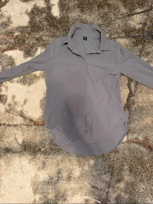 Ljusgrå skjorta med krage - En stilren ljusgrå skjorta med klassisk krage och långa ärmar. Skjortan har en enkel design utan synliga knappar framtill och är tillverkad i ett mjukt material som känns bekvämt mot huden. Perfekt för dig som gillar minimalistisk stil. Helt ny aldrig använd