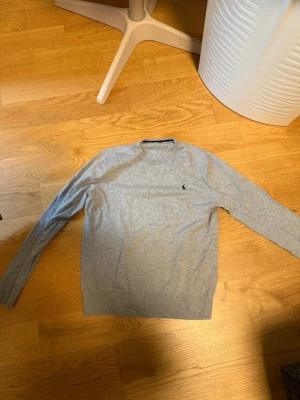 Grå tröja från Ralph Lauren - Tagg har tagits av av tidigare brukare! Snygg grå tröja från Ralph Lauren med klassisk rund halsringning och broderad logga på bröstet. Tröjan har långa ärmar och ribbade muddar vid ärmslut och nederkant. Perfekt för en stilren och enkel look.
