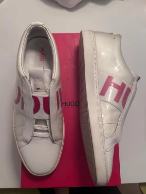 Vita sneakers från Hugo Boss - Snygga vita sneakers från Hugo Boss med breda elastiska band över vristen och stora röda bokstäver som stavar HUGO på sidan. Skorna har en chunky sula och är tillverkade i en mix av läder och textil. Perfekta för dig som gillar en clean och modern look. Nypris ca 2000, pris kan diskuteras.