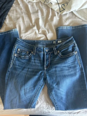 Blå bootcut jeans från Miss Me - Snygga blå bootcut jeans från Miss Me med kontrasterande vita sömmar och dekorativa detaljer på fickorna. Jeansen har klassisk femficksdesign och är tillverkade i denim med en cool tvätt. Perfekta för dig som gillar en avslappnad men trendig stil. Midja 39cm & innerbenslängd 82cm!