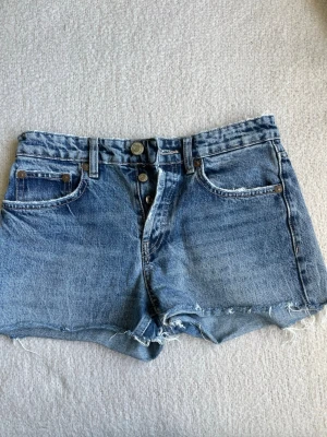 Blå denimshorts från Zara  - Säljer ett par klassiska blå jeansshorts. Shortsen har fem fickor, knappgylf och bälteshällor. Perfekta för sommaren och ger en avslappnad vibe till din outfit.