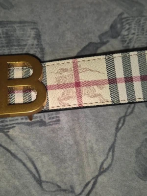 Rutigt bälte med guldigt B-spänne - Stilrent bälte med klassiskt rutigt mönster i beige, svart och rött. Bältet har ett stort guldigt B-format spänne och är perfekt för att lyfta vilken outfit som helst. Materialet är troligtvis syntet eller bomull.