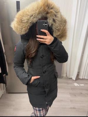 Svart parkas med pälskantad huva - Snygg svart parkas från Canada Goose med stor huva kantad av beige fuskpäls. Jackan har knäppning framtill, två stora fickor och en klassisk logga på ärmen. Perfekt för kalla dagar och riktigt varm.