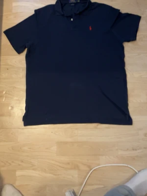 Mörkblå pikétröja från Ralph Lauren - Klassisk mörkblå pikétröja från Ralph Lauren med röd broderad logga på bröstet. Tröjan har krage, korta ärmar och knäppning framtill. Perfekt för en stilren och avslappnad look.