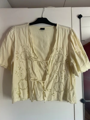 Gul blus med broderi Gina Tricot - Söt gul blus från Gina Tricot med broderade blommor och knytning framtill. Blusen har korta ärmar och en djup v-ringning. Tillverkad i ett lätt, strukturerat tyg som ger en avslappnad och bohemisk känsla.