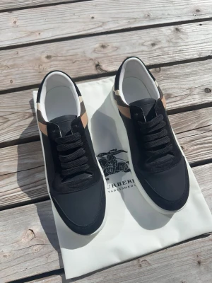 Svarta Burberry sneakers med rutdetalj - Snygga svarta sneakers från Burberry med klassisk beige och svart rutdetalj vid hälen. Skorna har svarta snören, vit sula och är tillverkade i en mix av läder och textil. Perfekta för dig som vill ha en stilren och trendig look.