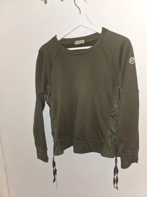 Olivgrön sweatshirt från Moncler - Cool orginal olivgrön sweatshirt från Moncler med långa ärmar och ribbade muddar. Snygga detaljer i form av glansiga sidopaneler och knytband nedtill. Moncler-logga på ena ärmen. Perfekt för en avslappnad men trendig look.