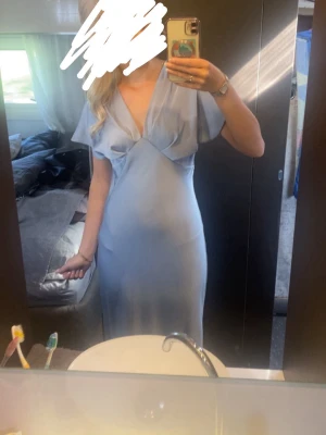 Ljusblå långklänning med v-ringning - Snygg ljusblå långklänning med v-ringad hals och korta ärmar. Klänningen har en elegant och feminin passform med mjukt fallande satin tyg, perfekt för sommarens tillfällen eller bal!. Stilren och enkel design som känns både modern och klassisk.
