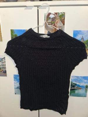 Svart stickad topp från Zara - Svart stickad topp från Zara med korta ärmar och ett diskret hålmönster. Toppen har rund halsringning och är figurnära i modellen. Perfekt att styla med jeans eller kjol för en trendig look.  