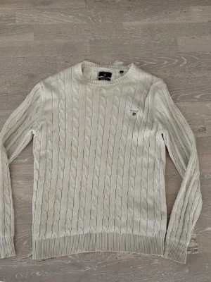Vit kabelstickad tröja från Gant - Kabelstickad tröja i vitt från Gant med rund halsringning och broderad logotyp på bröstet. Tillverkad i premium bomull, har ribbade muddar vid ärmslut och nederkant. Perfekt för en klassisk och stilren look.