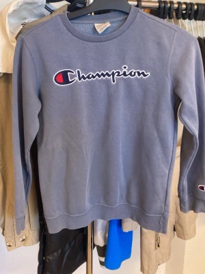 Blå långärmad Champion t-shirt - Säljer en blå långärmad t-shirt från Champion med det klassiska logotrycket i vitt, svart och rött på bröstet. Tröjan har rund halsringning och en liten Champion-logga på ärmen. Perfekt för en avslappnad och sportig stil.