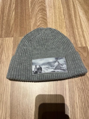Grå ribbstickad mössa från Revenite (företaget finns ej!) - Snygg grå ribbstickad mössa med uppvikt kant och en patch framtill som visar ett svartvitt motiv av två personer och ett berg. Perfekt accessoar för kalla dagar och ger en cool outdoor-vibe.