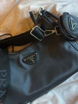 Svart axelväska från Prada nylon - Snygg svart axelväska från Prada i nylon med ikonisk trekantslogga i metall framtill. Väskan har bred axelrem med extra liten avtagbar ficka och silverfärgade detaljer. Perfekt för dig som gillar stilrena och trendiga accessoarer.