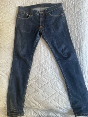 Mörkblå slim fit jeans från Nudie - Snygga mörkblå slim fit jeans från Nudie med klassiska fem fickor och orangea sömmar. Jeansen har en smal passform och är tillverkade i stretchig denim för extra komfort. Bakfickorna har Nudies ikoniska broderi.