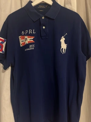 Blå pikétröja Polo Ralph Lauren XL - Blå pikétröja från Polo Ralph Lauren med broderad logga på bröstet, flaggdetaljer och tryck på ärmen. Klassisk krage med knappar och custom fit-passform. Perfekt för dig som gillar sportig och stilren design.