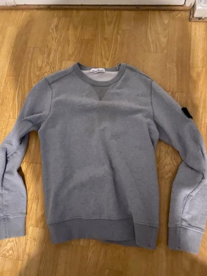 Grå sweatshirt från Stone Island - Grå sweatshirt från Stone Island med klassisk rund halsringning och ribbade muddar. Tröjan har den ikoniska Stone Island-loggan på vänster ärm och är tillverkad i mjuk bomull. Perfekt för en avslappnad och stilren look. Obs en liten fläck som knappts syns går ej bort i tvätten 