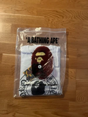 Vit A Bathing Ape t-shirt - Snygg vit t-shirt från A Bathing Ape med det klassiska stora aptrycket framtill och coola grafiska detaljer. T-shirten har rund hals och korta ärmar. Tillverkad i mjuk bomull och levereras i originalförpackning med taggar kvar.