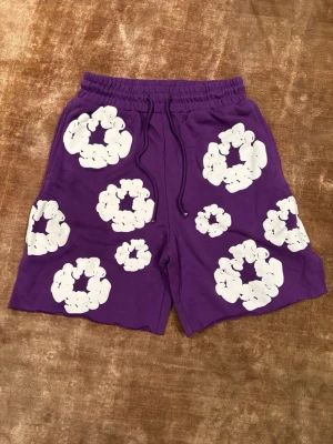 Lila Denim Tears Shorts M - Säljer ett par lila shorts med elastisk midja och snörning. Shortsen har stora vita blommiga mönster över hela tyget och en bakficka. Perfekta för sommaren och riktigt sköna att bära. Pris kan diskuteras 🙌🙌
