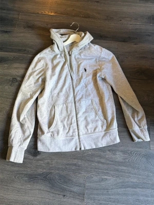 Ljusgrå hoodie från Polo Ralph Lauren - Snygg ljusgrå hoodie med dragkedja från Polo Ralph Lauren. Klassisk design med huva, två fickor framtill och broderad logga på bröstet. Perfekt för en avslappnad och stilren look.