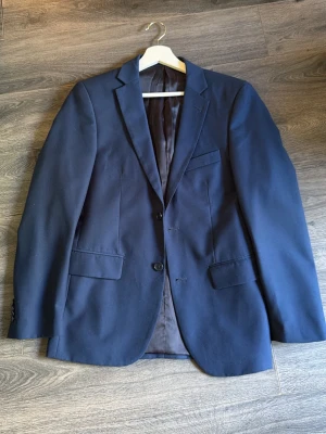 Mörkblå klassisk kavaj - Snygg mörkblå kavaj med två knappar framtill, klassisk passform och tre fickor. Fodrad insida och slits baktill. Perfekt för en stilren look till både skjorta och t-shirt.