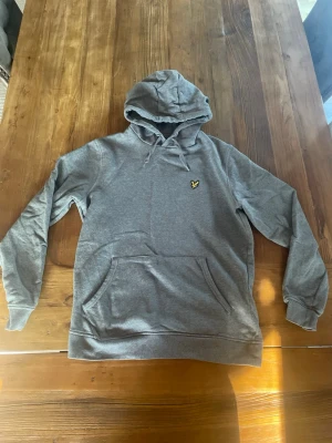 Grå hoodie från Lyle & Scott - Snygg grå hoodie från Lyle & Scott med klassisk gul broderad logga på bröstet. Hoodien har huva magficka och ribbade muddar vid ärmslut och nederkant. Perfekt för en avslappnad och stilren look.