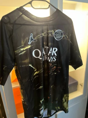 PSG x Jordan svart matchtröja - Säljer en svart PSG x Jordan matchtröja med coolt mönster i grönt och gult. Tröjan har korta ärmar, Dri-Fit-material och Paris Saint-Germain-logga på bröstet. Perfekt för fotboll eller träning. Qatar Airways sponsortryck på framsidan.