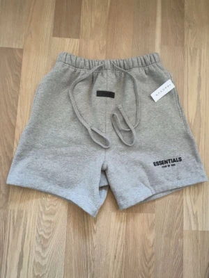 Grå shorts från Essentials Fear of God - Gråa shorts från Essentials Fear of God med elastisk midja och snörning. Mjukt material och logga framtill. Perfekta för en avslappnad stil och passar till många olika outfits.