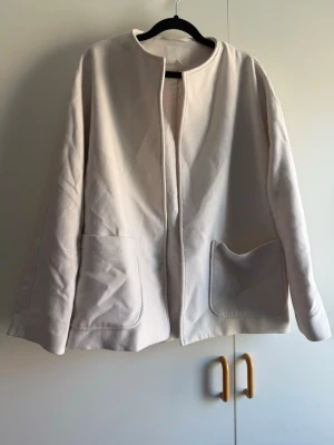 Vit cardiganjacka med fickor - Stilren beige cardiganjacka med rund halsringning och två stora framfickor. Jackan har en rak och loose passform samt långa ärmar. Materialet ser ut att vara bomull eller en bomullsblandning, vilket gör den mjuk och bekväm att bära. Perfekt för lager på lager och enkel att matcha. Köpt begagnad och endast provad. Katt finns i hem!