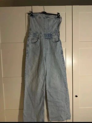 Ljusblå denim jumpsuit  - Snygg ljusblå denim jumpsuit med bandeau topp och raka, vida ben. Jumpsuiten har klassiska jeansdetaljer som fickor fram och bak samt bälteshällor. Tillverkad i denim med snygga sömmar som framhäver midjan.  Finns en ytters liten fläck på benet (se på sista bilden), annars som ny. Knappt använd 💗✨Storlek L, men är lite mindre, samt tightare runt bröstet om man har lite större/större bröst, bröstkorg  Jag är 172cm