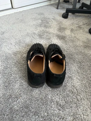 Svarta sneakers med nitar från Louboutin - Säljer ett par svarta sneakers från Christian Louboutin med coola svarta nitar på tån. Skorna har snörning, är låga och har en ovandel i mocka. Insidan är beige och sulan är svart. Perfekta för dig som vill sticka ut med din stil.