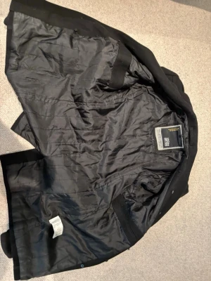 Svart jacka från Jack & Jones - Stilren svart jacka från Jack & Jones med slät yta och diskret design. Jackan har helsvart foder och klassisk passform, perfekt för dig som gillar enkel och tidlös stil. Märket syns på insidan och materialet känns mjukt och följsamt.