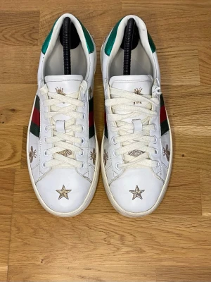 GC vita sneakers med brodyr - GC sneakers i vitt läder med gröna och röda sidoremsor samt guldbroderade bin och stjärnor. Klassisk låg modell med vita snören och grön detalj på hälen. Snygg och unik design som sticker ut.