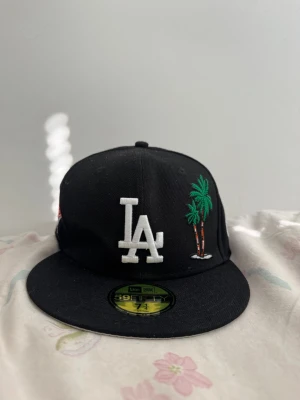 Svart LA Dodgers keps New Era 59Fifty - Säljer en svart LA Dodgers keps från New Era 59Fifty med broderad vit LA-logga och palmer framtill. På sidan finns ett färgglatt 1988 World Series-märke. Klassisk flat brim och skön passform, perfekt för dig som gillar streetwear och sportig stil.