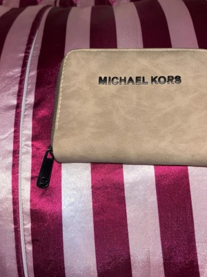 Beige plånbok från Michael Kors - Snygg beige plånbok från Michael Kors med silverfärgad logga på framsidan. Plånboken har dragkedja runt och är tillverkad i ett slätt material som ger en stilren look. Perfekt storlek för att få plats med kort och kontanter.